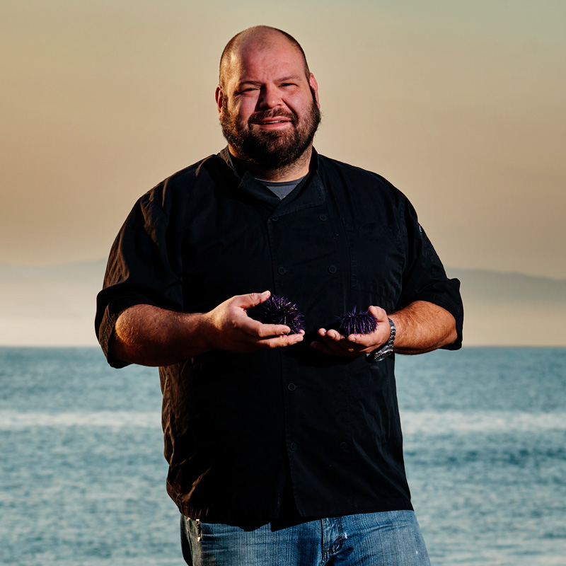 Chef Paul Grossi holding a pile of purple sea urchins on a beach